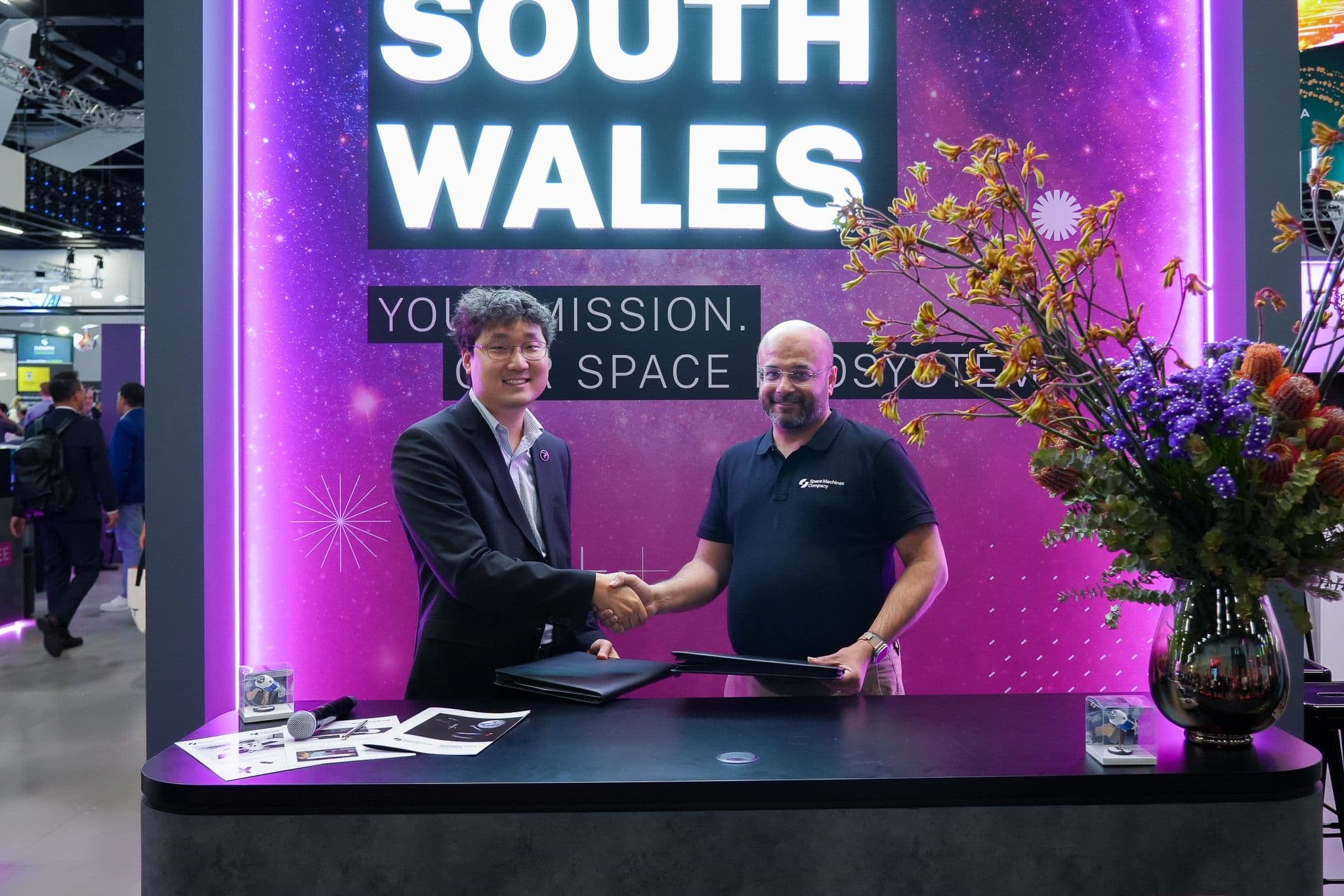 Space Machines Company and Perigee Aerospace representatives shaking hands at the partnership signing, beside a photo of a Kickstage propulsion engine.